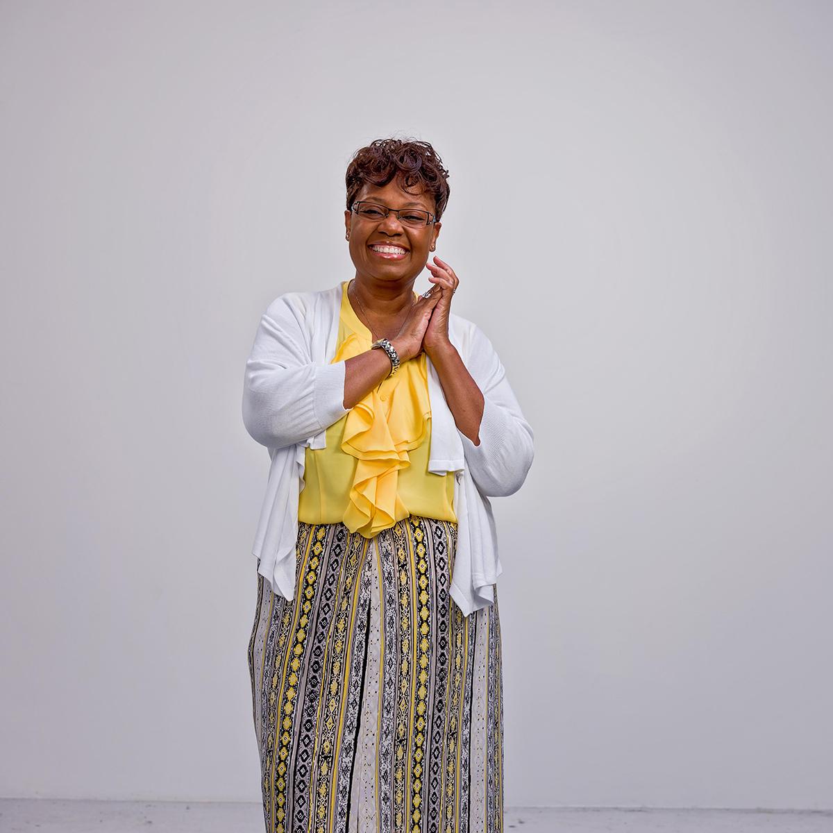 Photo of Rita Armstrong, a smiling Black woman with glasses and short hair, wearing a yellow blouse and colorful skirt