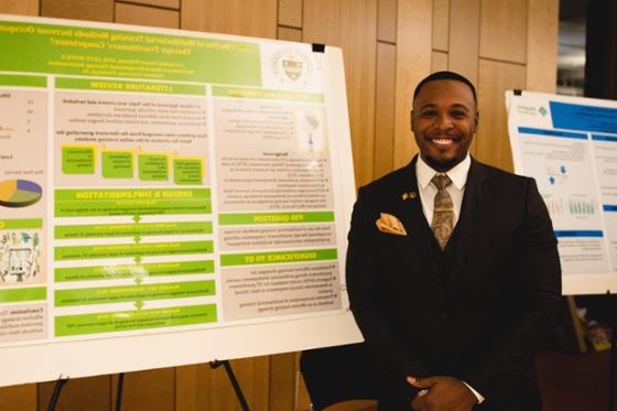 Photo of a smiling man in black suit and tie, posing in front of poster presentation. 