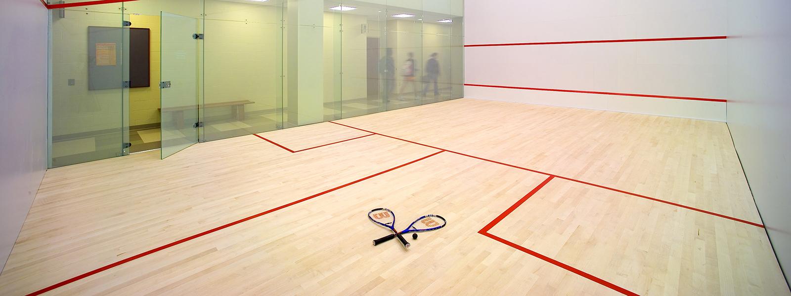 Photo of a squash court in the gym, with a ball and two rackets on the ground. 
