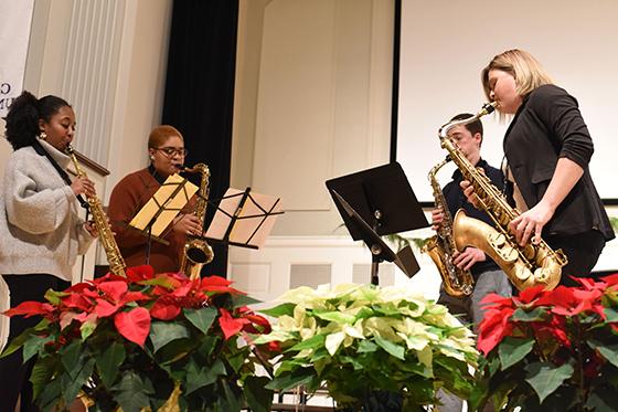 Photo of four brass players performing on a stage surrounded by red poinsettias. 