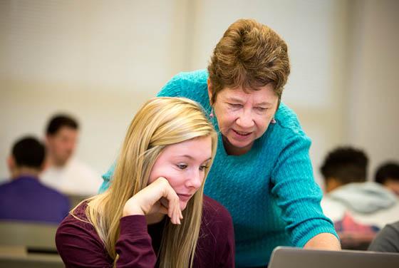图为一名年长的女教员俯身看着一名波胆网站的学生，指着一台电脑