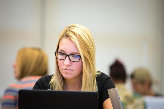 Photo of a white blonde woman in glasses working on a computer in a classroom