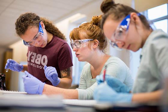Photo of three female Chatham University students in protective goggles and gloves working in a lab