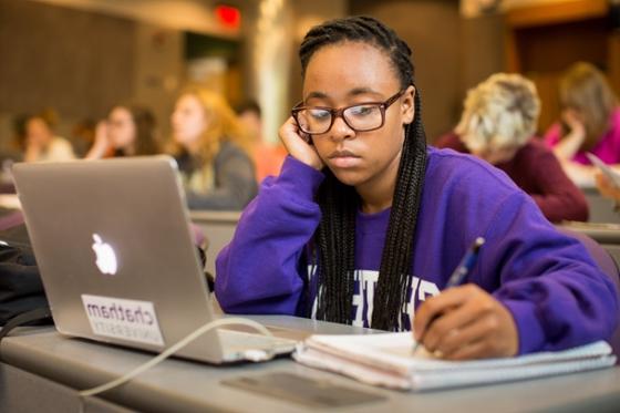 Photo of a student in a Chatham University sweatshirt, working at a laptop in a lecture hall and taking notes